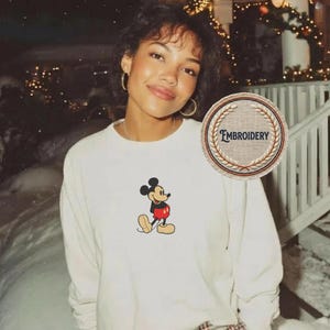 May include: Cream-coloured long-sleeve shirt with an embroidered Mickey Mouse design. The cartoon character is shown in his classic outfit with red shorts and yellow shoes. The shirt has a crew neck and a relaxed fit.