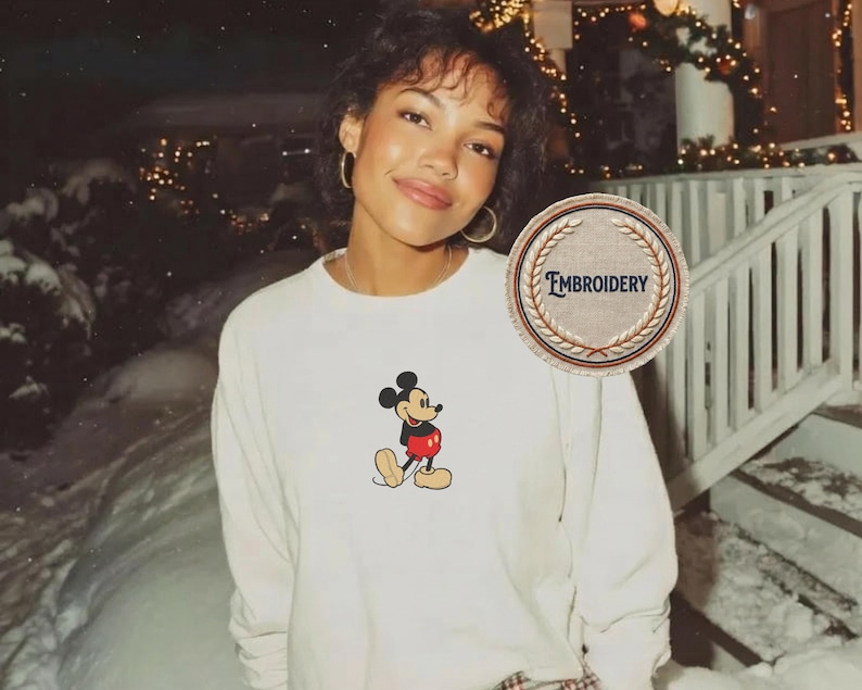 May include: Cream-coloured long-sleeve shirt with an embroidered Mickey Mouse design. The cartoon character is shown in his classic outfit with red shorts and yellow shoes. The shirt has a crew neck and a relaxed fit.