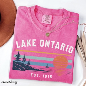 May include: A bright pink short-sleeve t-shirt featuring the text LAKE ONTARIO and EST. 1615 in white. The graphic includes a dark blue shoreline with pine trees, a small sailboat, and a sunset over horizontal yellow, teal, and blue stripes.