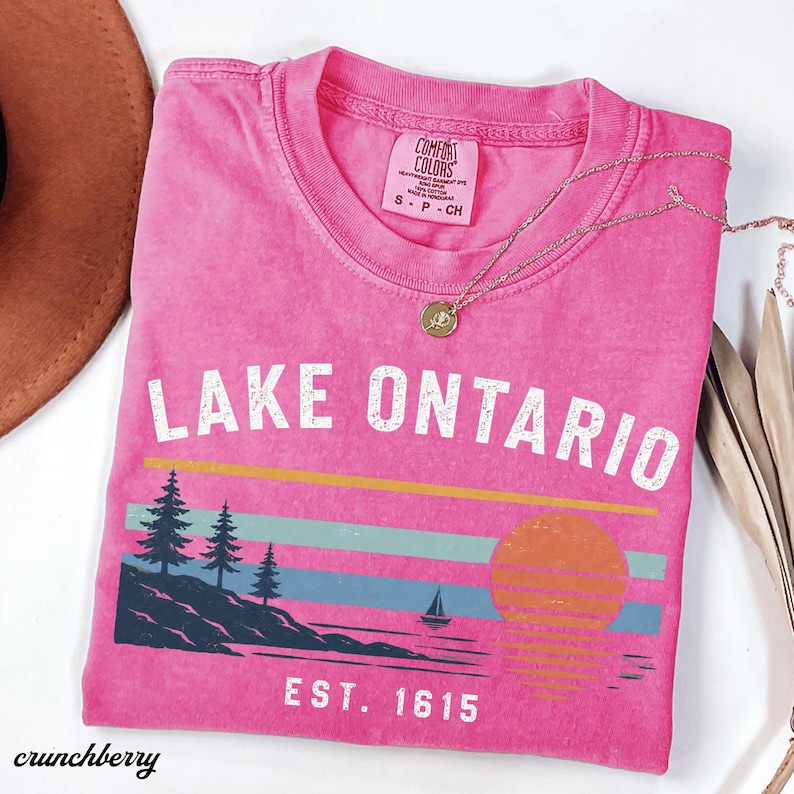 May include: A bright pink short-sleeve t-shirt featuring the text LAKE ONTARIO and EST. 1615 in white. The graphic includes a dark blue shoreline with pine trees, a small sailboat, and a sunset over horizontal yellow, teal, and blue stripes.