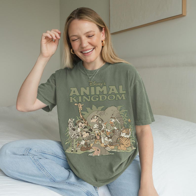 May include: Olive green t-shirt featuring the text "Disney's Animal Kingdom" above a graphic of various cartoon animals. The graphic includes a zebra, giraffe, elephant, and tiger. The shirt is a relaxed fit.
