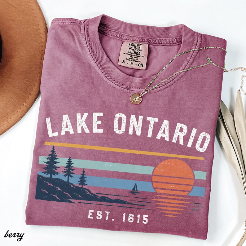 May include: A berry-colored t-shirt featuring the white text LAKE ONTARIO and EST. 1615. The graphic includes a landscape with pine trees, a sailboat, and a setting sun over water, rendered in horizontal stripes of yellow, teal, and blue.