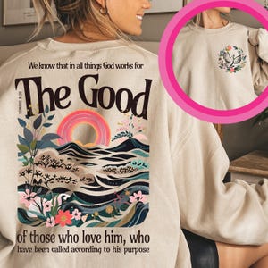 May include: A beige sweatshirt with a colorful graphic design featuring a rainbow, waves, and flowers. The text "We know that in all things God works for The Good of those who love him, who have been called according to his purpose" is printed on the front of the sweatshirt.
