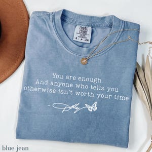 May include: A blue jean coloured short-sleeve t-shirt with a crew neckline. The shirt features white text that reads: You are enough. And anyone who tells you otherwise isn't worth your time - Dolly, accompanied by a small white butterfly graphic.
