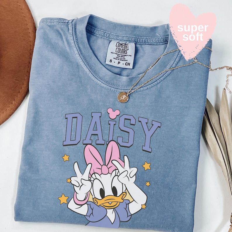 Puede incluir: Camiseta azul Comfort Colors con un gr&aacute;fico de Daisy Duck y la palabra "DAISY" en morado. Daisy hace el signo de la paz. Tambi&eacute;n se ven un collar dorado y una pegatina rosa en forma de coraz&oacute;n. Talla S.