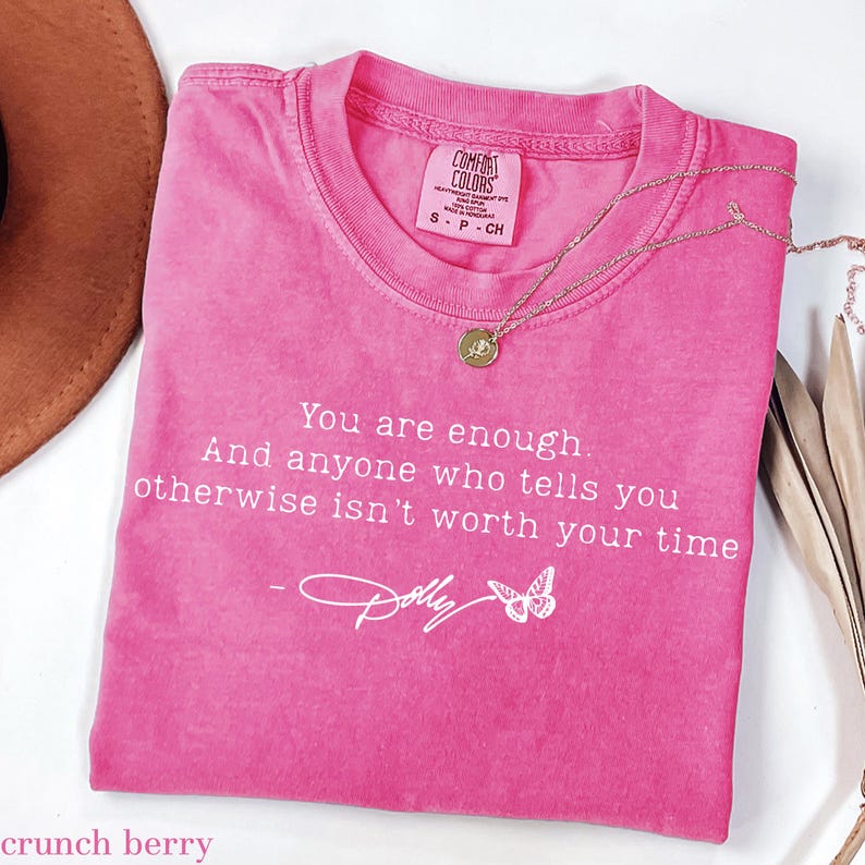 May include: A pink short-sleeved t-shirt with a crew neckline and a Comfort Colors label. The shirt features white text that reads: You are enough. And anyone who tells you otherwise isn't worth your time - Dolly, with a small butterfly graphic.