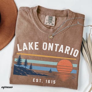 May include: A brown short-sleeve t-shirt with a graphic print featuring the text LAKE ONTARIO and EST. 1615. The design includes a landscape with pine trees, a sailboat, and a sun over water in shades of orange, light blue, and dark blue.