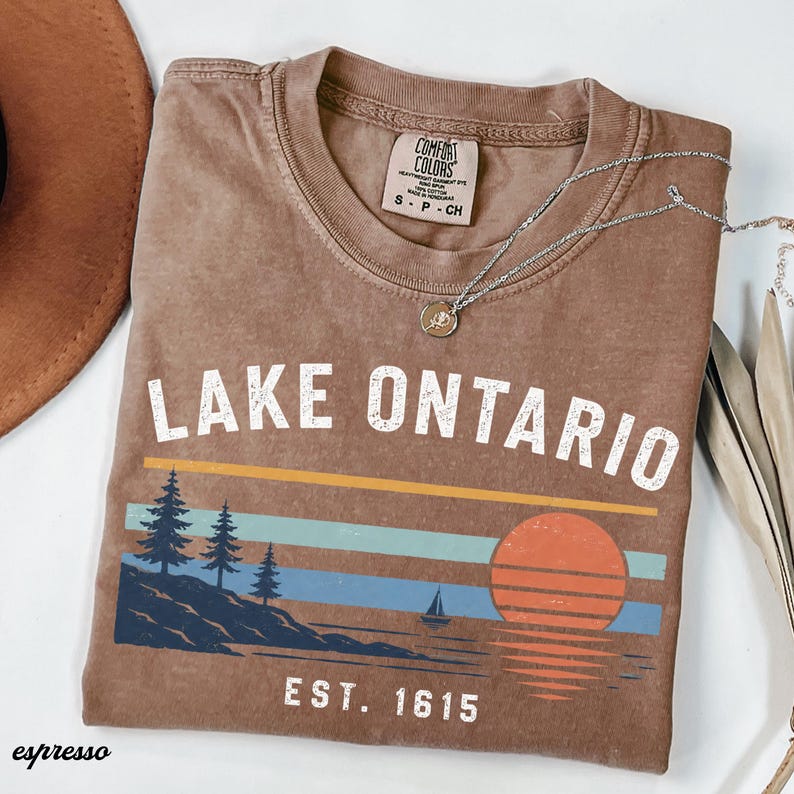 May include: A brown short-sleeve t-shirt with a graphic print featuring the text LAKE ONTARIO and EST. 1615. The design includes a landscape with pine trees, a sailboat, and a sun over water in shades of orange, light blue, and dark blue.