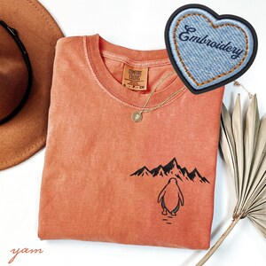 May include: Coral-colored t-shirt with a crew neck and a penguin graphic. The graphic features a penguin standing in front of a mountain range. A denim heart-shaped patch with the word "Embroidery" is also visible. A gold necklace and a brown hat are also in the image.