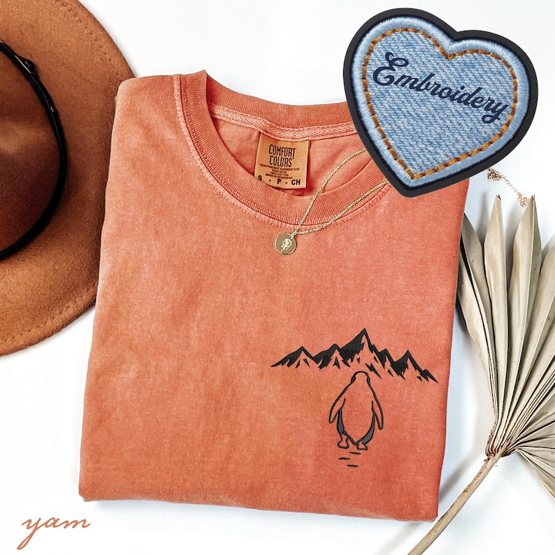 May include: Coral-colored t-shirt with a crew neck and a penguin graphic. The graphic features a penguin standing in front of a mountain range. A denim heart-shaped patch with the word "Embroidery" is also visible. A gold necklace and a brown hat are also in the image.