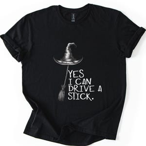 以下が含まれることがあります： 黒いTシャツ。魔女の帽子とほうきのグラフィック。白い文字で「YES I CAN DRIVE A STICK」と書かれています。ハロウィンやテーマのあるイベントに最適な、快適なトップスです。