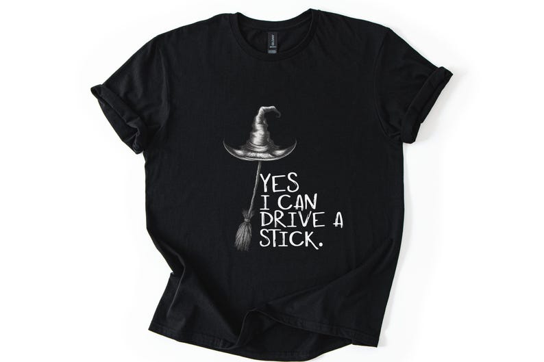 以下が含まれることがあります： 黒いTシャツ。魔女の帽子とほうきのグラフィック。白い文字で「YES I CAN DRIVE A STICK」と書かれています。ハロウィンやテーマのあるイベントに最適な、快適なトップスです。
