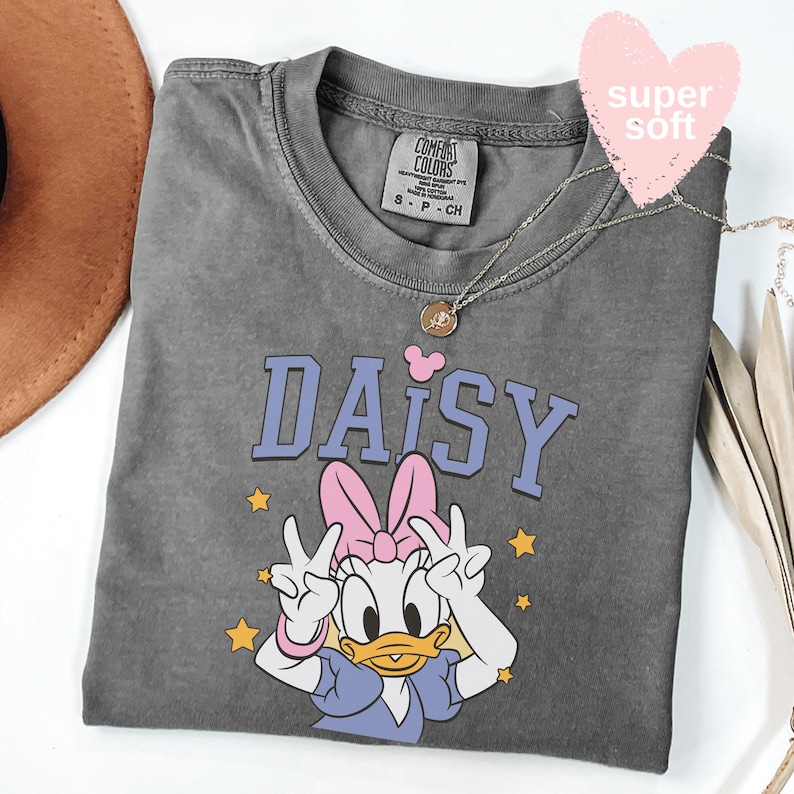 Puede incluir: Una camiseta gris Comfort Colors con un gr&aacute;fico de Daisy Duck y la palabra "DAISY" en letras azules. Daisy hace el signo de la paz, lleva un lazo rosa y est&aacute; rodeada de estrellas amarillas. Tambi&eacute;n se ve un collar plateado y una etiqueta rosa en forma de coraz&oacute;n.