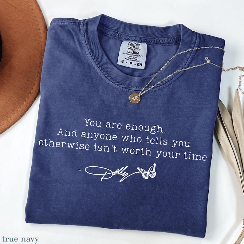 May include: A navy blue short-sleeve t-shirt with a white graphic print that reads: You are enough. And anyone who tells you otherwise isn't worth your time. The text includes a signature that reads Dolly and a small butterfly illustration.