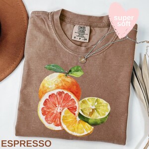 Op de afbeelding: Espresso-kleurig T-shirt met een aquarel illustratie van citrusvruchten, waaronder een grapefruit, een sinaasappel, een citroen en een limoen. Het shirt heeft een ronde hals en korte mouwen. Een zilveren ketting met een kleine hanger is over het shirt gedrapeerd. De woorden "super soft" staan op een roze hart.