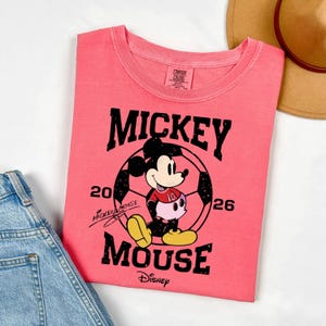 Puede incluir: Camiseta de color coral con un gr&aacute;fico de Mickey Mouse con uniforme de f&uacute;tbol, con el texto "Mickey Mouse" y "2026". La camiseta incluye el logotipo de Disney y una firma. Un sombrero color canela y unos vaqueros azules est&aacute;n en el fondo.