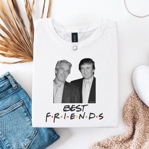 May include: White t-shirt with a black and white image of two men, and the words "BEST FRIENDS" in colourful letters. The t-shirt is folded on a white surface, with blue jeans and a decorative plant visible in the background.