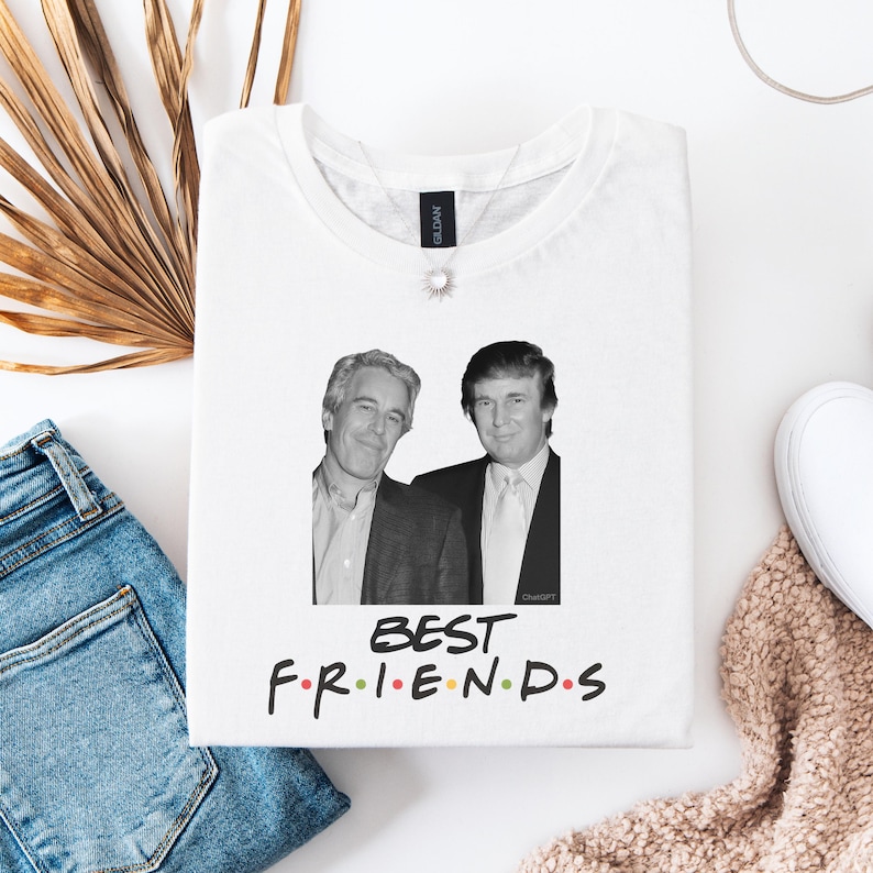 May include: White t-shirt with a black and white image of two men, and the words "BEST FRIENDS" in colourful letters. The t-shirt is folded on a white surface, with blue jeans and a decorative plant visible in the background.