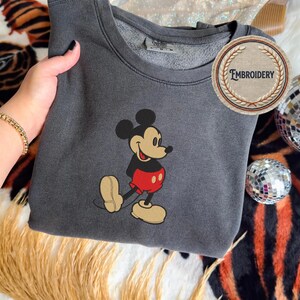 May include: A charcoal grey sweatshirt with an embroidered Mickey Mouse design. The cartoon mouse is wearing red shorts, a black top, and tan shoes. The sweatshirt has a crew neck and a circular "Embroidery" logo.