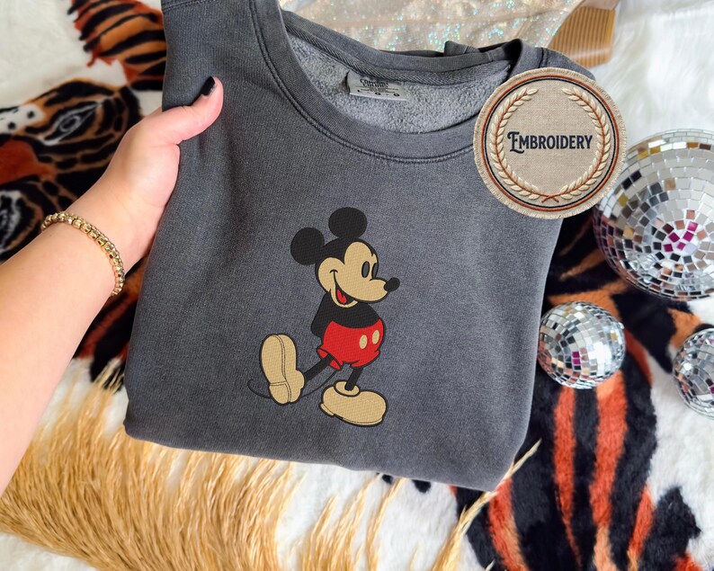 May include: A charcoal grey sweatshirt with an embroidered Mickey Mouse design. The cartoon mouse is wearing red shorts, a black top, and tan shoes. The sweatshirt has a crew neck and a circular "Embroidery" logo.