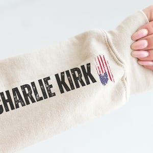 以下が含まれることがあります： ベージュ色のスウェットシャツの袖に、黒色のかすれた文字で「CHARLIE KIRK」とプリントされています。アメリカ国旗のグラフィックが袖の上にあります。背景は白です。