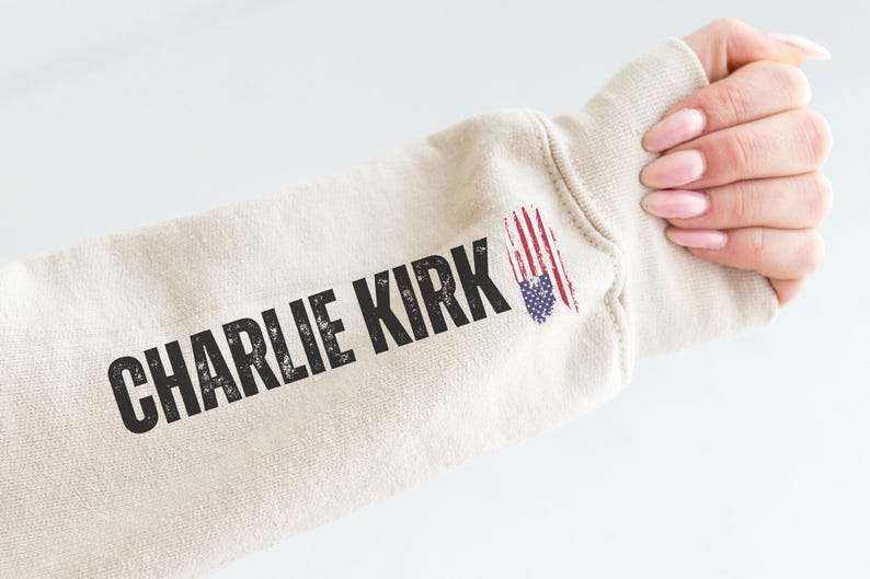 以下が含まれることがあります： ベージュ色のスウェットシャツの袖に、黒色のかすれた文字で「CHARLIE KIRK」とプリントされています。アメリカ国旗のグラフィックが袖の上にあります。背景は白です。