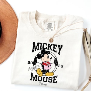 Puede incluir: Camiseta color crema con un gr&aacute;fico de Mickey Mouse en un bal&oacute;n de f&uacute;tbol. El dise&ntilde;o incluye el texto "Mickey Mouse" y el logotipo de Disney. Tambi&eacute;n se ven un collar dorado y un sombrero marr&oacute;n.