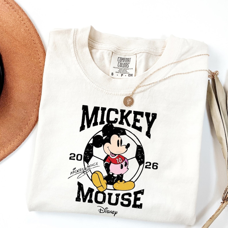 Puede incluir: Camiseta color crema con un gr&aacute;fico de Mickey Mouse en un bal&oacute;n de f&uacute;tbol. El dise&ntilde;o incluye el texto "Mickey Mouse" y el logotipo de Disney. Tambi&eacute;n se ven un collar dorado y un sombrero marr&oacute;n.