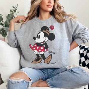 May include: A grey sweatshirt with a Minnie Mouse design. The cartoon character is shown in a red polka-dot dress, black shoes, and a flower on her head. The Disney logo is printed below the character.