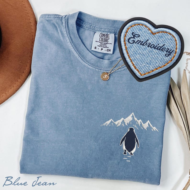 May include: Blue jean colored t-shirt with an embroidered penguin and mountain scene. A denim heart-shaped patch with the word "Embroidery" is visible. A gold necklace with a pendant is also present.