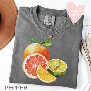 Op de afbeelding: Een grijs Comfort Colors T-shirt met een aquarel illustratie van citrusvruchten, waaronder een grapefruit, sinaasappel en limoen. Het shirt heeft een ronde hals en korte mouwen. Een zilveren ketting met een hanger is over het shirt gedrapeerd. Het woord "PEPPER" staat onderaan.