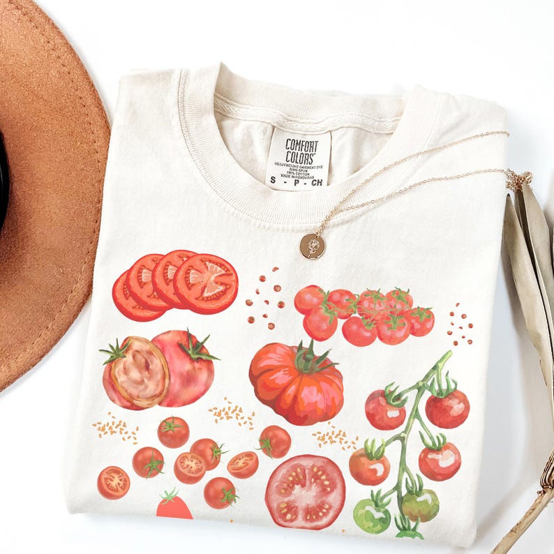 May include: A white t-shirt with a red tomato print. The print features whole tomatoes, sliced tomatoes, and cherry tomatoes. The shirt has a tag that says "Comfort Colors".