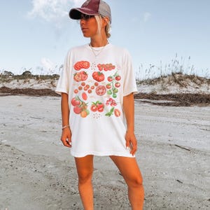 May include: A white oversized t-shirt with a graphic print of red tomatoes, green leaves, and a few red berries. The shirt is worn by a person on a sandy beach.