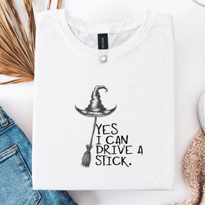 以下が含まれることがあります： 魔女の帽子とほうきのグラフィックが描かれた白いTシャツ。「YES I CAN DRIVE A STICK」の文字がグラフィックの下にプリントされています。青いジーンズ、白いスニーカー、ネックレスと合わせてコーディネートされています。
