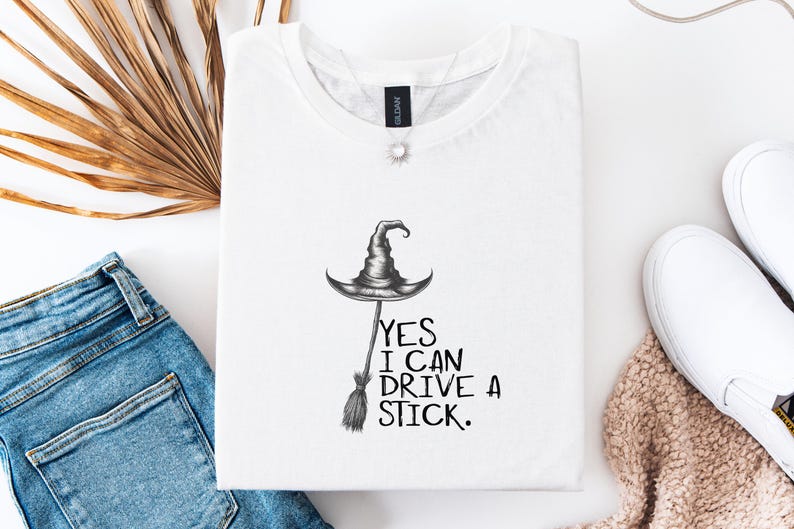 以下が含まれることがあります： 魔女の帽子とほうきのグラフィックが描かれた白いTシャツ。「YES I CAN DRIVE A STICK」の文字がグラフィックの下にプリントされています。青いジーンズ、白いスニーカー、ネックレスと合わせてコーディネートされています。