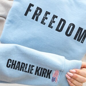 以下が含まれることがあります： 薄い青色のスウェットシャツ。黒の擦り切れたフォントで「FREEDOM」の文字がプリントされています。袖には「CHARLIE KIRK」の文字と小さなアメリカ国旗のピンが付いています。スウェットシャツはニュートラルカラーの表面にあります。