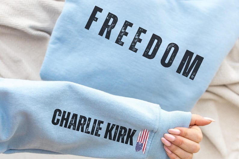 以下が含まれることがあります： 薄い青色のスウェットシャツ。黒の擦り切れたフォントで「FREEDOM」の文字がプリントされています。袖には「CHARLIE KIRK」の文字と小さなアメリカ国旗のピンが付いています。スウェットシャツはニュートラルカラーの表面にあります。