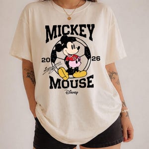 Puede incluir: Camiseta color crema con un dise&ntilde;o gr&aacute;fico de Mickey Mouse en un bal&oacute;n de f&uacute;tbol. El texto incluye "MICKEY MOUSE" y "20 26" con el logo de Disney. Mickey lleva una camiseta roja con el n&uacute;mero 10.