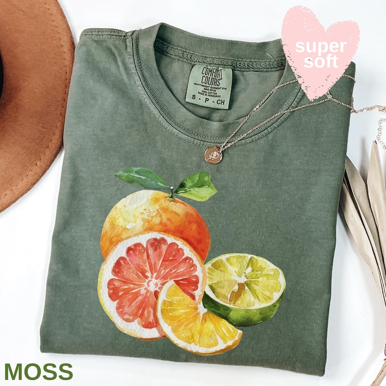 Op de afbeelding: Mosgroen T-shirt met een aquarel illustratie van citrusvruchten, waaronder een sinaasappel, grapefruit, limoen en sinaasappelschijf. Het shirt heeft een ronde hals en korte mouwen. Een delicate gouden ketting ligt op het shirt.