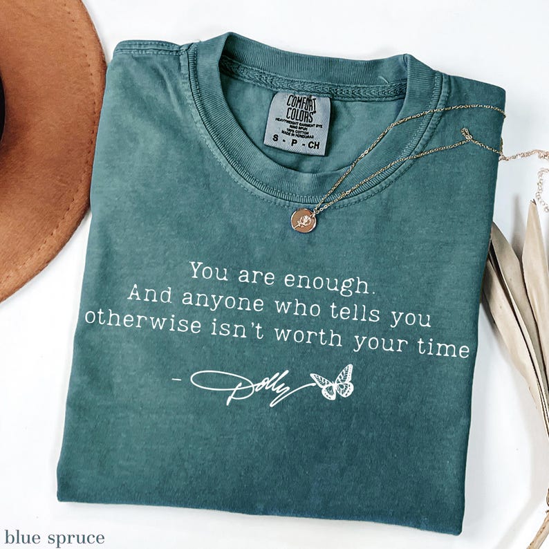May include: A blue spruce coloured t-shirt with a crew neckline and a Comfort Colors brand label. The shirt features white text that reads: You are enough. And anyone who tells you otherwise isn't worth your time - Dolly, with a butterfly graphic.
