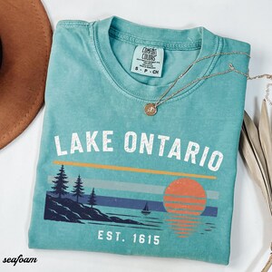 May include: A seafoam green short-sleeve t-shirt with a crew neck and a Comfort Colors label. The graphic features the text LAKE ONTARIO and EST. 1615, with a landscape illustration of pine trees, a sailboat, and a sunset over the water.