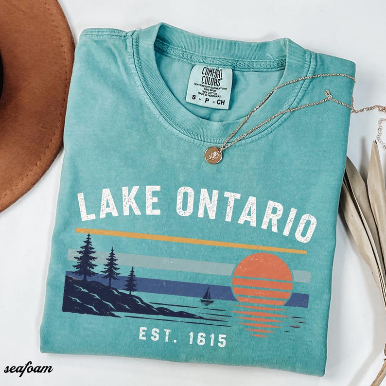 May include: A seafoam green short-sleeve t-shirt with a crew neck and a Comfort Colors label. The graphic features the text LAKE ONTARIO and EST. 1615, with a landscape illustration of pine trees, a sailboat, and a sunset over the water.