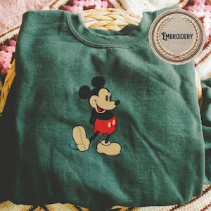 May include: A dark green sweatshirt with an embroidered Mickey Mouse design. Mickey is wearing his iconic red shorts, black top, and yellow shoes. The sweatshirt has a crew neck, and a circular patch with the word "Embroidery" is visible.
