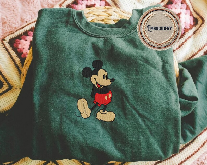 May include: A dark green sweatshirt with an embroidered Mickey Mouse design. Mickey is wearing his iconic red shorts, black top, and yellow shoes. The sweatshirt has a crew neck, and a circular patch with the word "Embroidery" is visible.