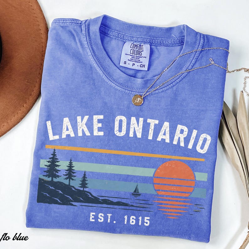 May include: A periwinkle blue short-sleeve t-shirt featuring a graphic print with the text LAKE ONTARIO and EST. 1615. The design includes a dark blue shoreline with pine trees, a small sailboat, and a large orange sun over horizontal stripes.