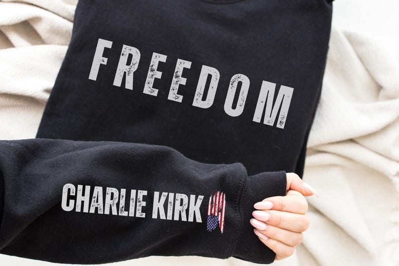以下が含まれることがあります： 「FREEDOM」の文字がかすれた白い文字でプリントされた黒いスウェットシャツ。袖には「CHARLIE KIRK」の文字と小さなアメリカ国旗のグラフィックが施されています。スウェットシャツはニュートラルカラーの生地の上に展示されています。
