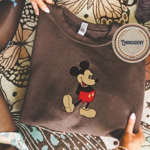 May include: A brown sweatshirt with an embroidered Mickey Mouse design. The cartoon mouse is wearing his iconic red shorts, black jacket, and yellow shoes. A circular patch with the word "EMBROIDERY" is visible.