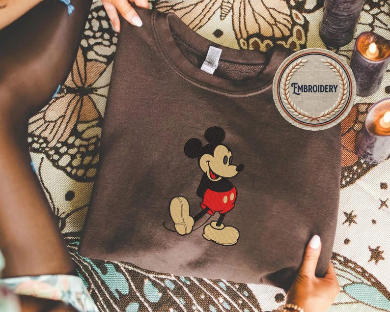 May include: A brown sweatshirt with an embroidered Mickey Mouse design. The cartoon mouse is wearing his iconic red shorts, black jacket, and yellow shoes. A circular patch with the word "EMBROIDERY" is visible.