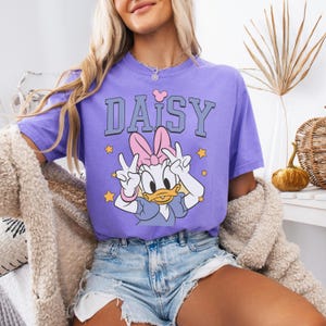 Puede incluir: Una camiseta lila con un dise&ntilde;o de Daisy Duck haciendo el signo de la paz. La palabra "DAISY" est&aacute; impresa encima del personaje en gris y blanco, con un lazo rosa y estrellas amarillas. La camiseta se combina con pantalones cortos vaqueros.