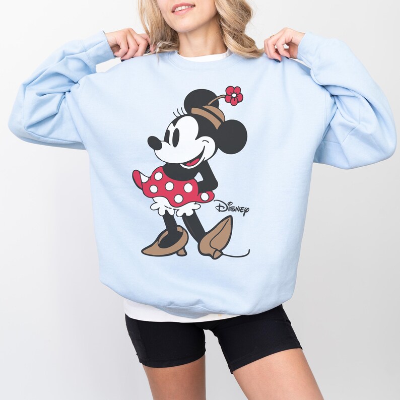 May include: Pale blue sweatshirt with a Minnie Mouse design. The illustration depicts Minnie Mouse in a red polka dot dress, brown shoes, and a flower on her head. The Disney logo is printed below the character.